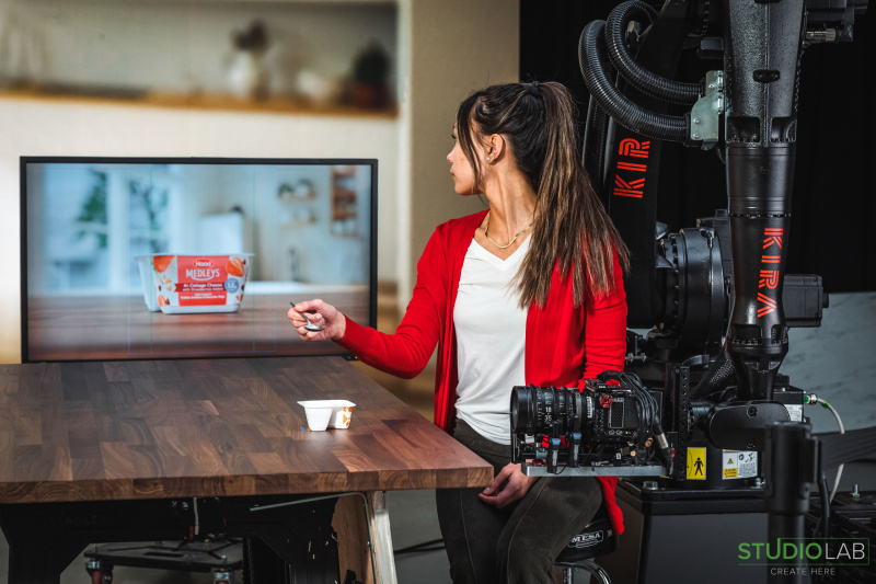 A woman in a red cardigan sits next to a camera rig with "KIRA" branding. She points at a monitor displaying Hood Medleys cottage cheese on a wooden table.