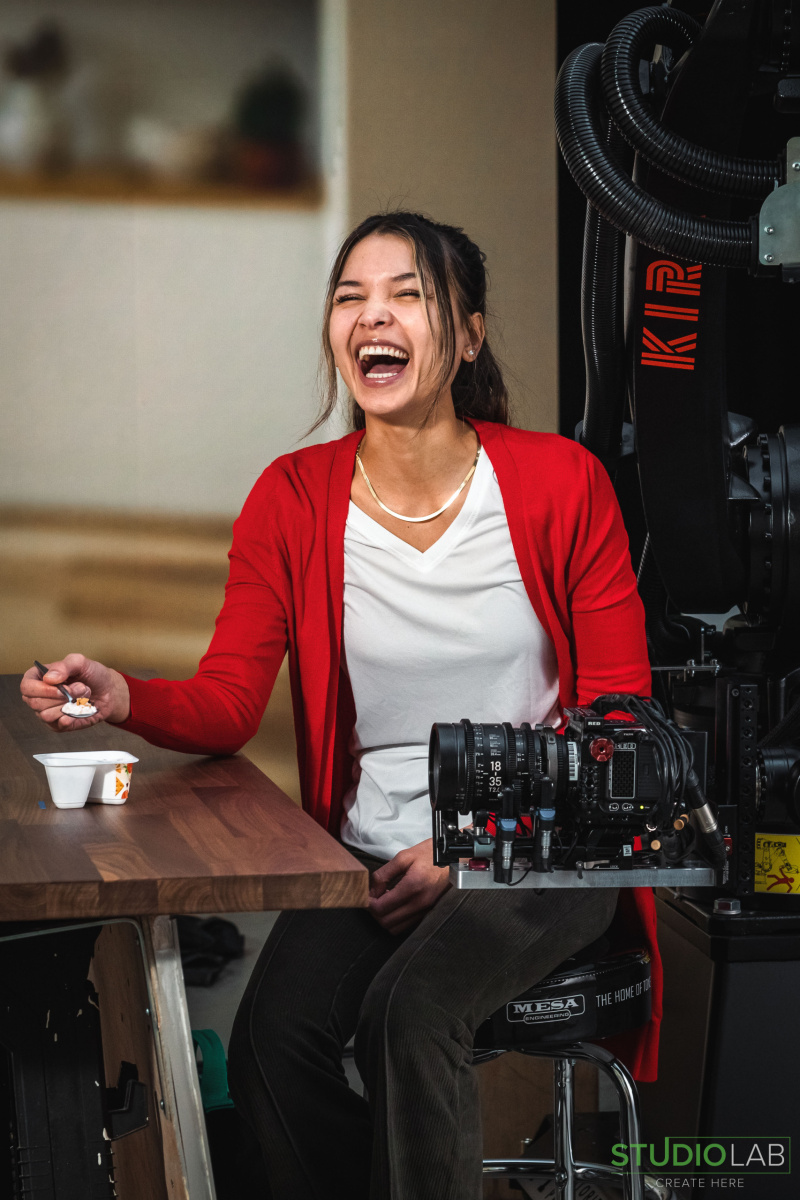 A woman dressed in red laughs during a scene on set while holding a spoon near a cottage cheese cup. A nearby camera rig is positioned, capturing the moment.