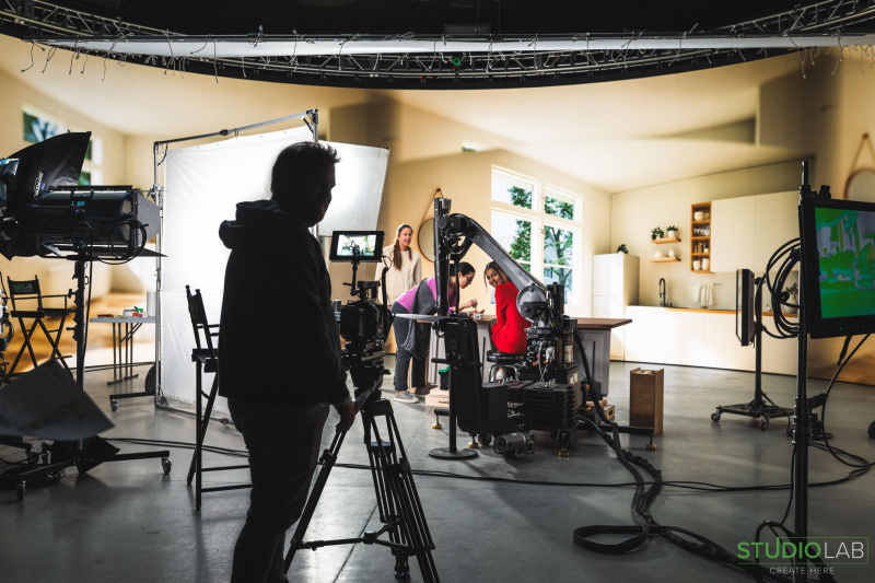 A camera crew sets up inside a bright kitchen set, with lighting and a robotic camera rig, preparing to film an actress in a red dress for the next scene.